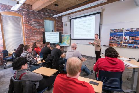 Professor standing in a classroom giving a lecture with a slide about securing federal systems in the background